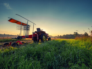 Old rusty tractor with grass cutting machine in a agriculture field with tall grass at stunning sunrise. Farming industry. Beautiful nature scene at special time. Nobody.