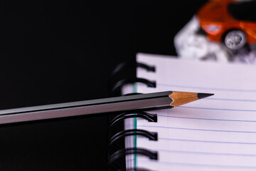 High angle close up shot of a pencil with spiral note book and red colored toy car blurred over...