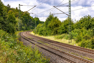 Two parallel tracks of a railroad line next to trees and catenary poles