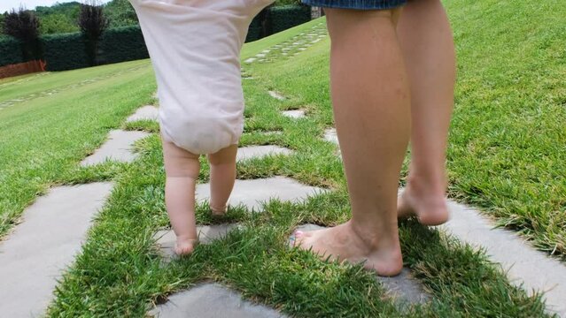 Young mother guiding her baby to walk in summer park, barefoot on the grass, slow motion