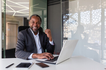 Portrait financial winner, mature african american boss smiling and looking at camera, businessman holding hand up in triumph gesture and happy with financial achievement results, man inside office.