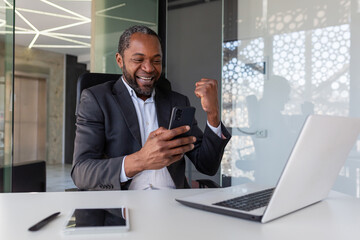 Mature successful african american businessman received online notification of winning message at workplace inside office, man holding phone, using app on smartphone, happy winner with achievement.