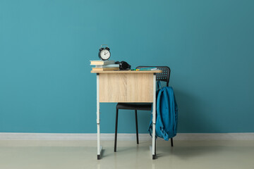 Modern school desk with backpack, alarm clock and stationery in room near blue wall