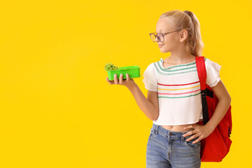 Happy girl in eyeglasses with backpack and lunchbox on yellow background - obrazy, fototapety, plakaty