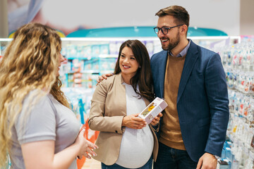 Attractive middle age couple enjoying in buying clothes and appliances for their new baby. Experienced saleswoman trying to help. Expecting baby concept. - obrazy, fototapety, plakaty