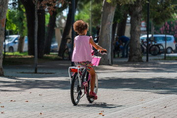 Cute pink-clad girl with curly hair in pink shirt rides pink bicycle, summer morning sunlight. Street lined with trees, cars blurred in background. Dry leaves and tree shadows on the road