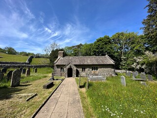 Fototapeta na wymiar St Leonard's Church, with a paved path, wild grass and flowers, hills and trees, set against a blue sky in, Chapel-le-Dale, UK