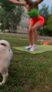 With her pug dog as a companion, a sportive young woman practices sports exercises amidst the vibrant backdrop of a summer park.