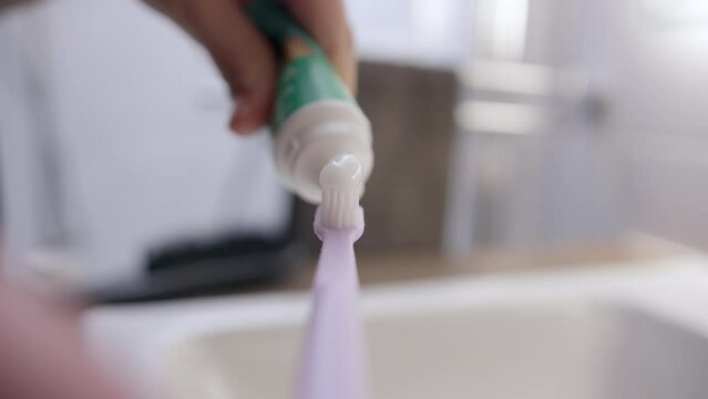 POV view of unrecognizable girl hand squeezing white toothpaste onto purple toothbrush and wets it with water. concept of maintaining personal hygiene. Morning or evening routine. 