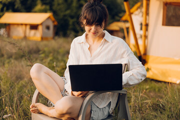 Beautiful woman in cozy outfit works at laptop while sitting on chair near the tent glamping