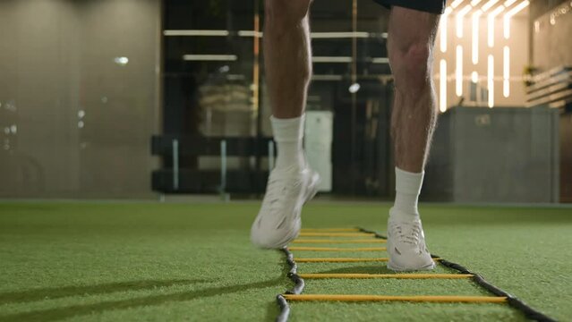 Unknown professional footballer running exercise jumping between ladder sticks male sportsman man soccer player muscular legs in sneakers fast jogging in sport gym training workout on football pitch