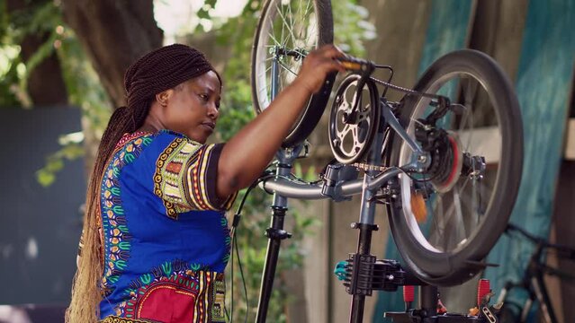 Passionate african american lady diligently examining and adjusting bike pedals and chains. Side-view portrait of smiling young black woman making annual adjustments to modern bicycle in yard.