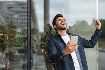 Handsome smiling indian man holding smartphone using mobile app, rejoicing