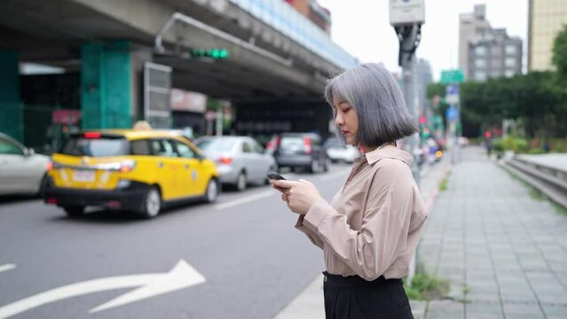 台湾台北の大通りでスマートフォンを操作しながらタクシーを待つ女性 A woman waits for a cab on a main street in Taipei, Taiwan, while operating a smartphone.