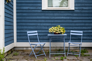 A purple-colored metal patio table near a blue wooden horizontal clapboard siding exterior wall. There's a flower pot with yellow flowers on the small square table. The ground is patio flat rocks.