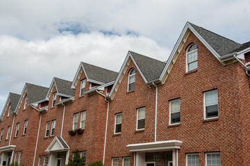 A row of brown brick townhouses with peaked gable roofs, and double hung windows with white trim. The shingles are black asphalt. The adjoined facade row houses are multi-story residential buildings. 