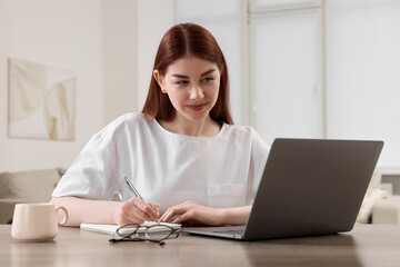 Beautiful woman using laptop and writing notes at wooden table indoors