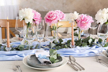 Beautiful table setting. Plate with greeting card, napkin and branch near glasses, peonies, burning candles and cutlery on table indoors