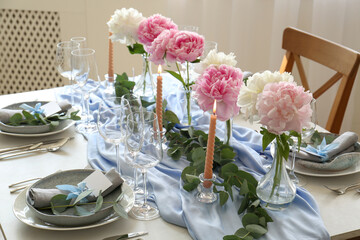 Beautiful table setting. Plates with greeting cards, napkins and branches near glasses, peonies, burning candles and cutlery on table indoors