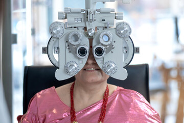 Middle-aged Indian or Nepali woman looking through Optical Phoropter during eye exam, diagnostic ophthalmology