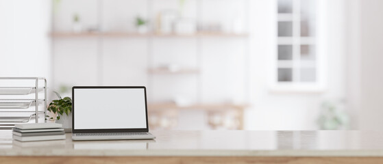 A white screen laptop mockup on a tabletop with a blurred modern white room in the background.