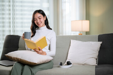 A happy Asian woman reading a book while having her morning coffee on a sofa in living room