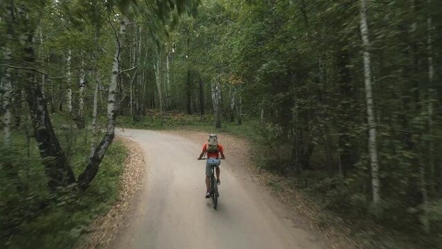A tourist cyclist in a helmet with a backpack walks along the road in a picturesque forest on a mountain bike. Shooting from a drone from the back