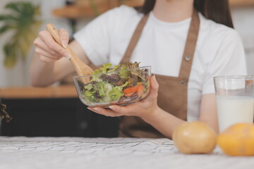 Delicious fruit and vegetables on a table and woman cooking. Housewife is cutting green cucumbers on a wooden board for making fresh salad in the kitchen.