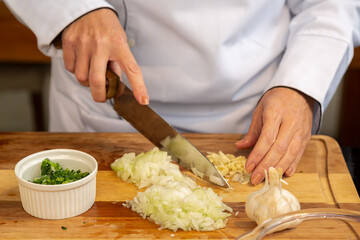 mãos de uma cozinheira usando Dolmã, preparando e picando alho e cebola sobre uma tabua de madeira e mais pote branco com salsa e cebolinha picados.