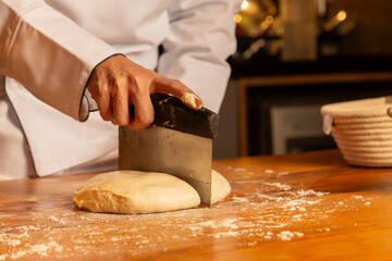 Detalhe das mãos de uma cozinheira profissional dividindo a massa crua de pão sobre a mesa de madeira coberta de farinha. - obrazy, fototapety, plakaty