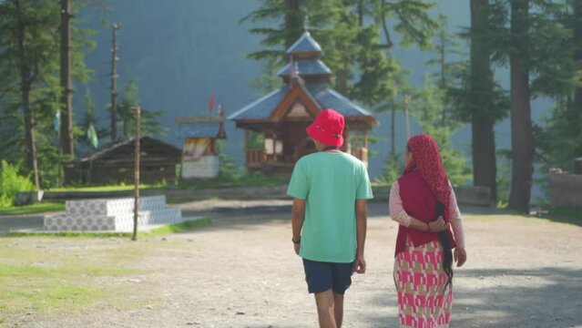 Jibhi, Himachal Pradesh, India - June 21 2023: Young Man and  woman walk mahadev temple in the meadow of Shahgarh, surrounded by Deodar Tree and Himalayas mountain in Sainj Valley.