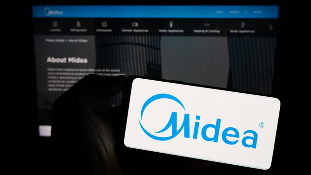 Stuttgart, Germany - 08-28-2023: Person holding smartphone with logo of Chinese home appliances company Midea Group on screen in front of website. Focus on phone display.