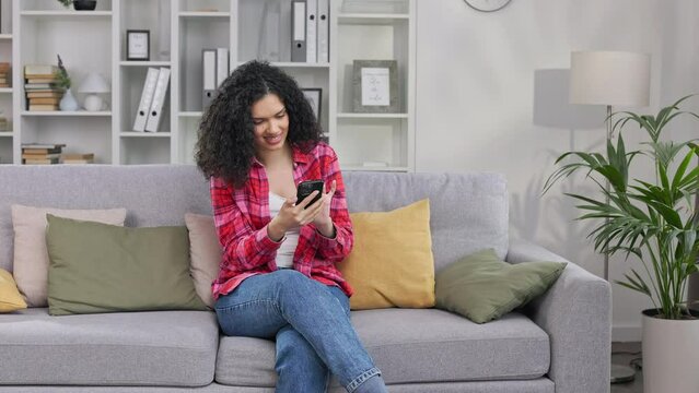 Smiling woman of african american ethnicity in blue jeans and colored shirt sitting on couch with smartphone and viewing pictures from vacation. Lady feeling happy when remembering of joyful days.