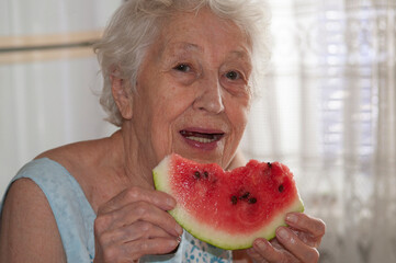 Smiling old senior woman holding a slice of fresh watermelon looking at camera at home background -...