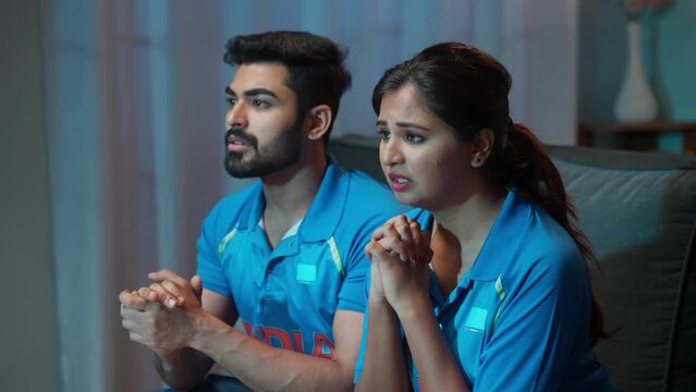 Couple with Indian jersey watching tensed cricket sports match at home during evening on tv - concept of nervous or Nail-biting Game and Emotional Reaction.