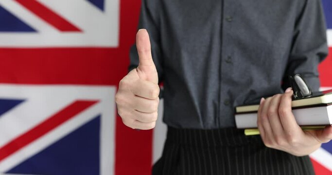 Female English teacher shows thumb up standing with books and glasses on background of Great Britain flag. Language learning and approval from specialist in school