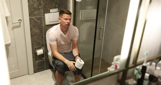 Bored man poops sitting on toilet bowl with toilet paper in hands in front of mirror in bathroom. Concept of natural process slow motion
