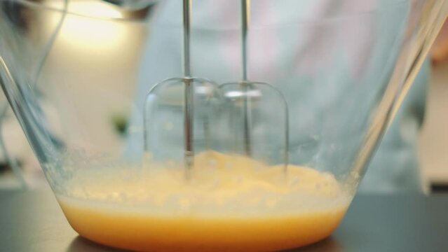 A woman beats eggs with a mixer into a strong foam in a glass plate. Close-up of the table. Cooking in the kitchen