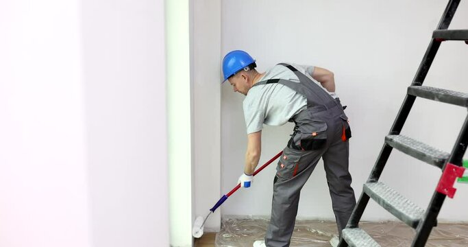 Builder in helmet and uniform overalls paints walls of studio apartment. Worker painting room using roller on long stick. House renovation slow motion