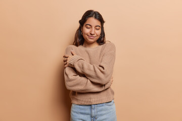 Pleased brunette Iranian woman daydreams embraces herself with both hands recalls romantic event keeps eyes closed smiles gently wears knitted jumper and jeans isolated over brown background.