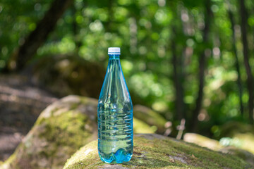 A transparent plastic bottle with clean drinking water stands on a large stone covered with moss in the middle of a forest