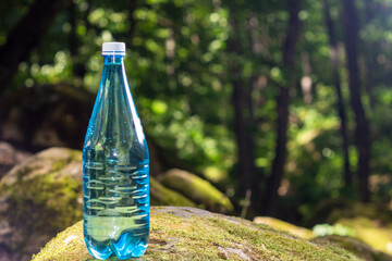 A transparent plastic bottle with clean drinking water stands on a large stone covered with moss in the middle of a forest