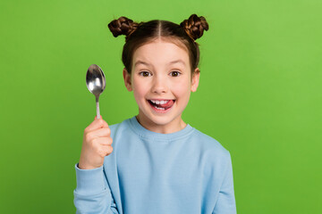 Portrait of little girl with brown buns hairstyle wear blue pullover lick teeth hold spoon wait...