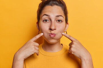 Careless young woman with hair bun points index fingers at cheeks holds breath looks at camera dressed in casual t shirt isolated over vivid yellow background tries to be funny. Human face expressions - obrazy, fototapety, plakaty
