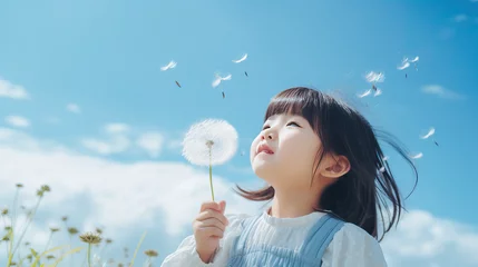 Foto op Plexiglas Photo of a little Asian girl blowing a dandelion, toddler child holding a dandelion flower on blue sky background, looking upwards, close-up summer portrait with copy space © Favebrush