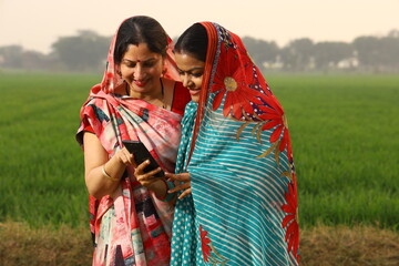 Happy rural Indian women standing in a mustard agricultural field and surfing through the mobile phone in their hand.