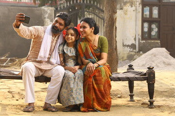 Happy Indian Rural family of Mother, father and daughter sitting together smiling on cot outside their home in front yard using mobile phone.