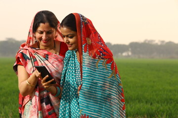 Happy rural Indian women standing in a mustard agricultural field and surfing through the mobile phone in their hand.