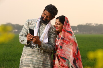 Happy rural Indian villager family of husband and wife sitting together outside their home/house/cottage Husband showing wife on the mobile phone.