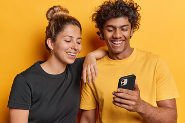 Positive young woman and man friends concentrated at smartphone with cheerful expressions read news online watch funny video together dressed in casual t shirts isolated over yellow background
